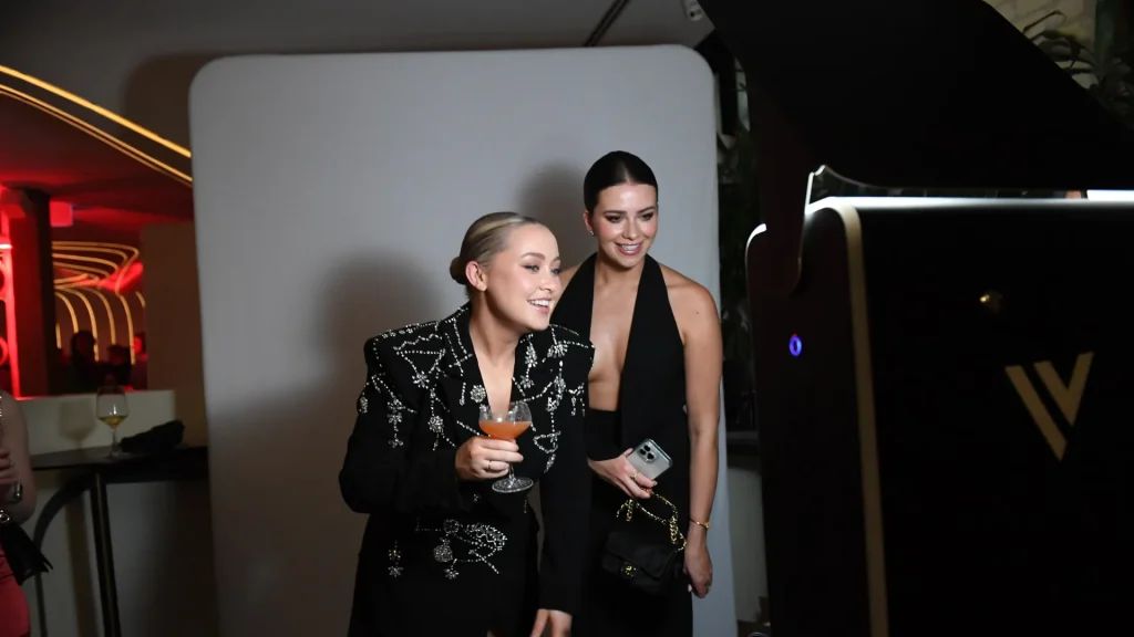 Branded photo booths. Best photo booth rentals in Toronto. Ineffable Views. Two women posing and smiling in front of a photo booth at an evening event, one holding a cocktail and the other holding a phone, with a white backdrop.
