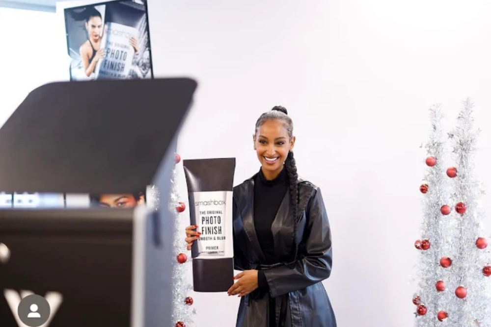 Branded photo booths. Best photo booth rentals in Toronto. Ineffable Views. Woman in black leather outfit smiling and holding oversized Smashbox Photo Finish primer prop at branded event with silver Christmas trees.